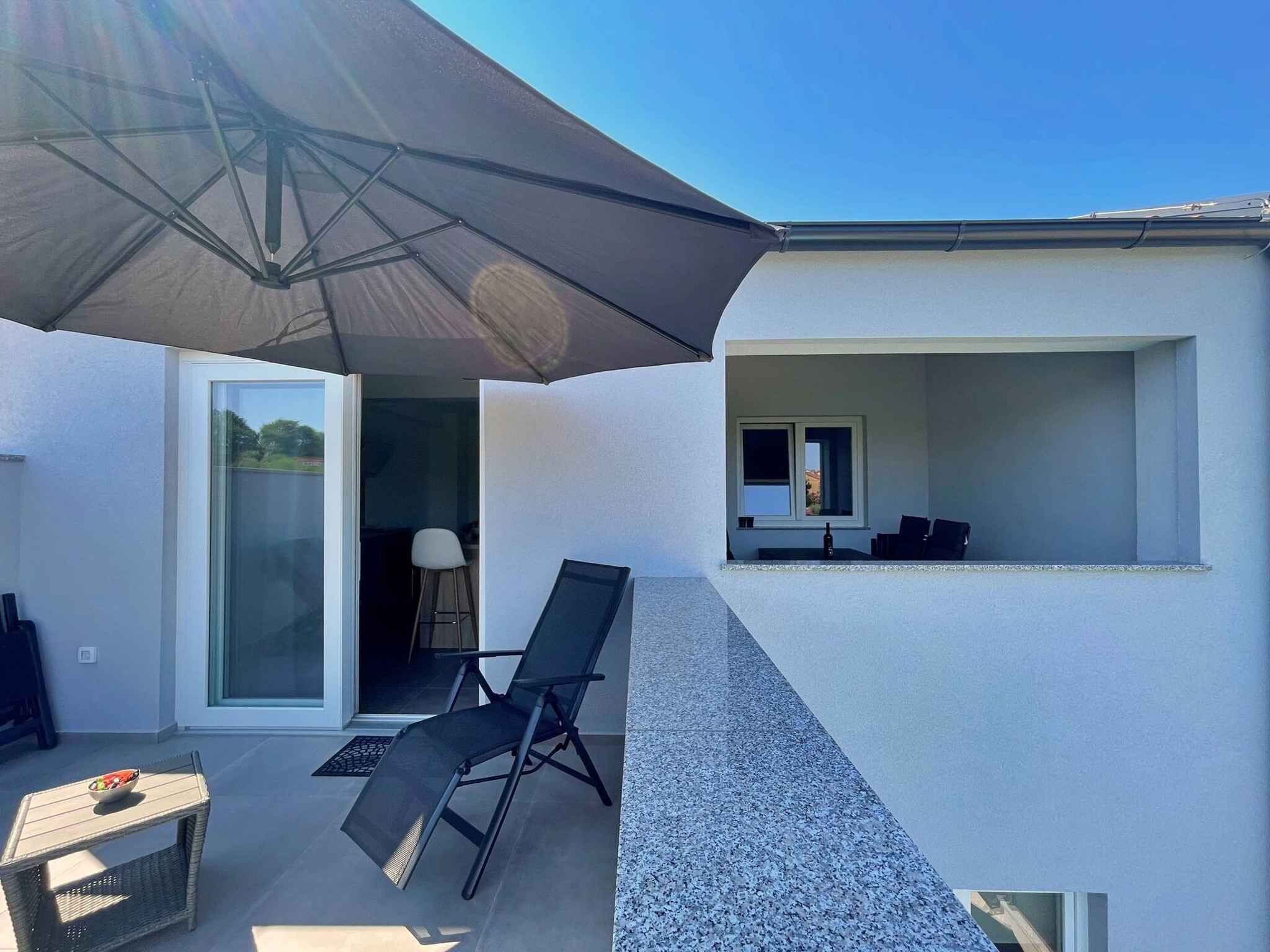 A modern outdoor patio with a glass door leading to a room, a black chair, a small table with a bowl, and a large umbrella.