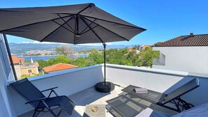 Eine Dachterrasse mit schwarzem Sonnenschirm, Liegestühlen, einem Glastisch und Blick auf einen See und Gebäude.