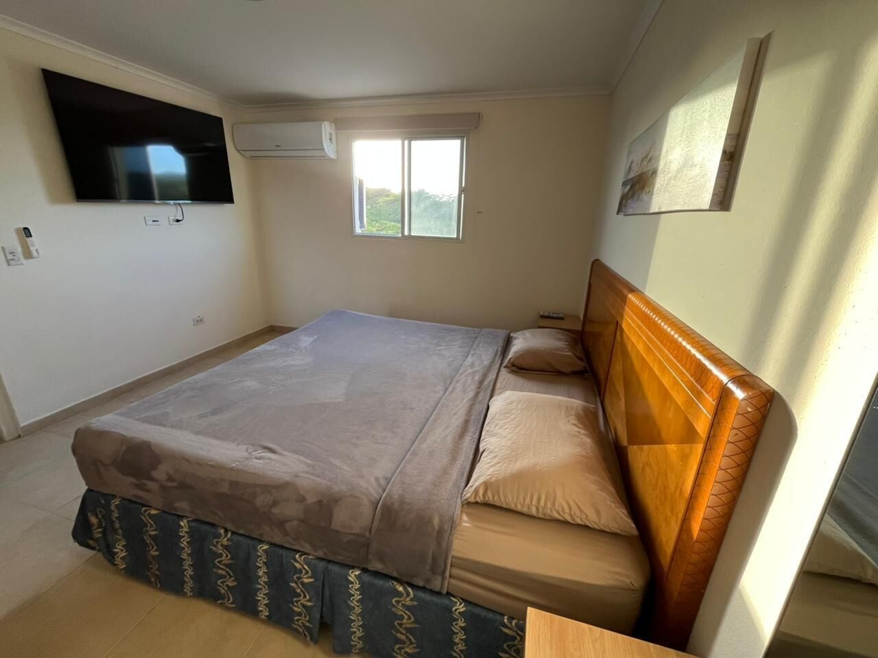 A bedroom with a bed, a TV mounted on the wall, a window, and a painting.