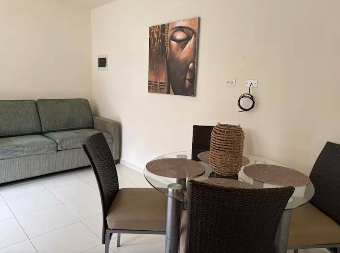 Un coin repas avec une table en verre entourée de chaises en osier, d’un canapé gris et d’une œuvre d’art accrochée au mur.