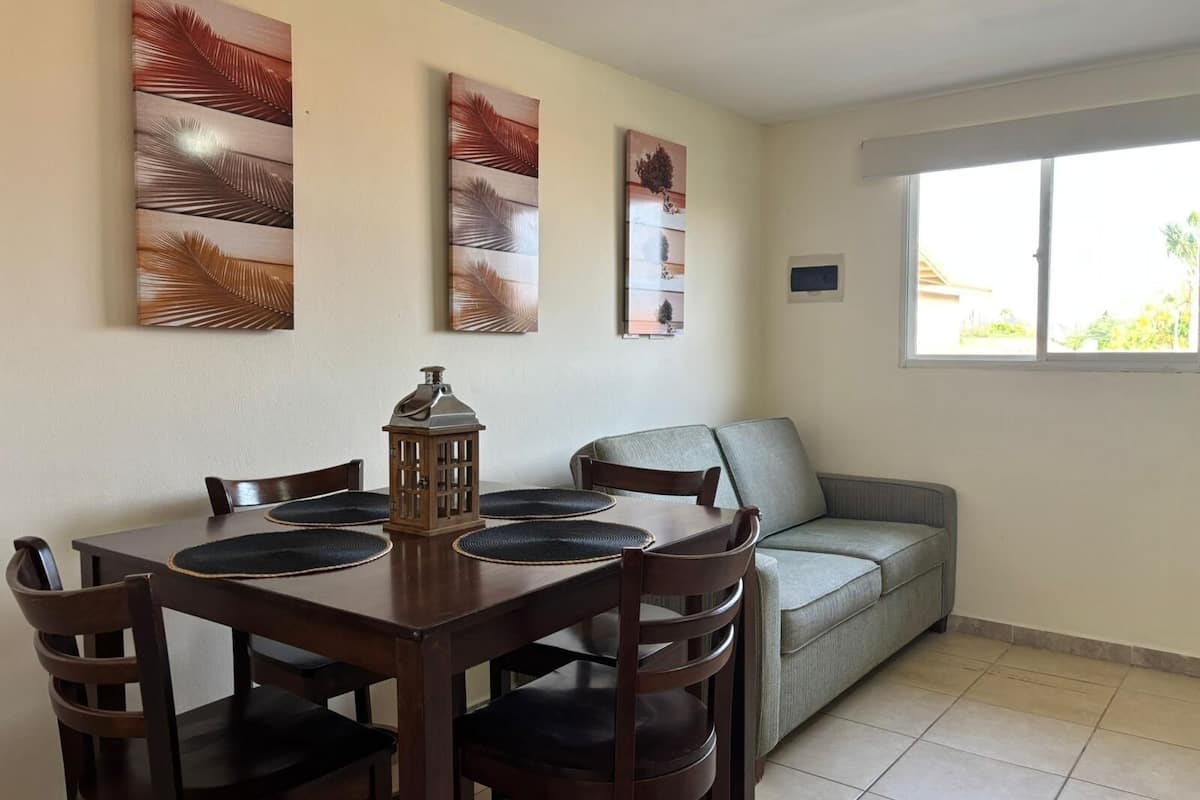 Un comedor con una mesa de madera, cuatro sillas y un farol decorativo. Un sofá gris está apoyado contra la pared, y hay tres obras de arte colgadas en la pared sobre la mesa.