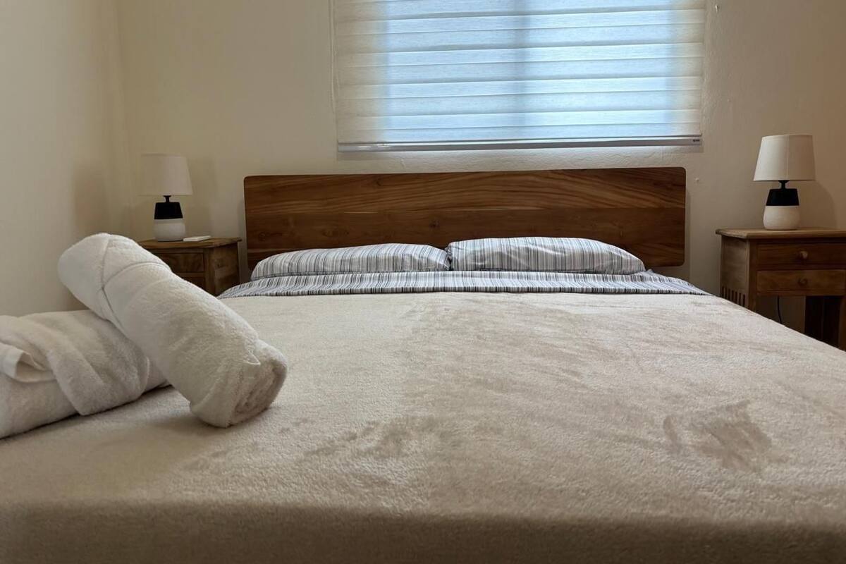 Una cama bien hecha con cabecera de madera, dos mesitas de noche con lámparas y una ventana con persianas.
