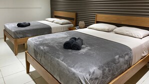 Habitación de hotel con dos camas, cada una con ropa de cama gris y cabeceros de madera. Entre las camas hay una mesita con una lámpara.