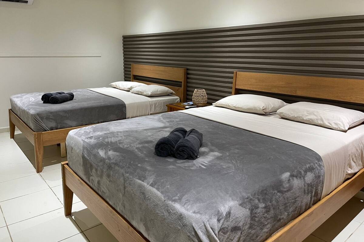 Habitación de hotel con dos camas, cada una con ropa de cama gris y cabeceros de madera. Entre las camas hay una mesita con una lámpara.