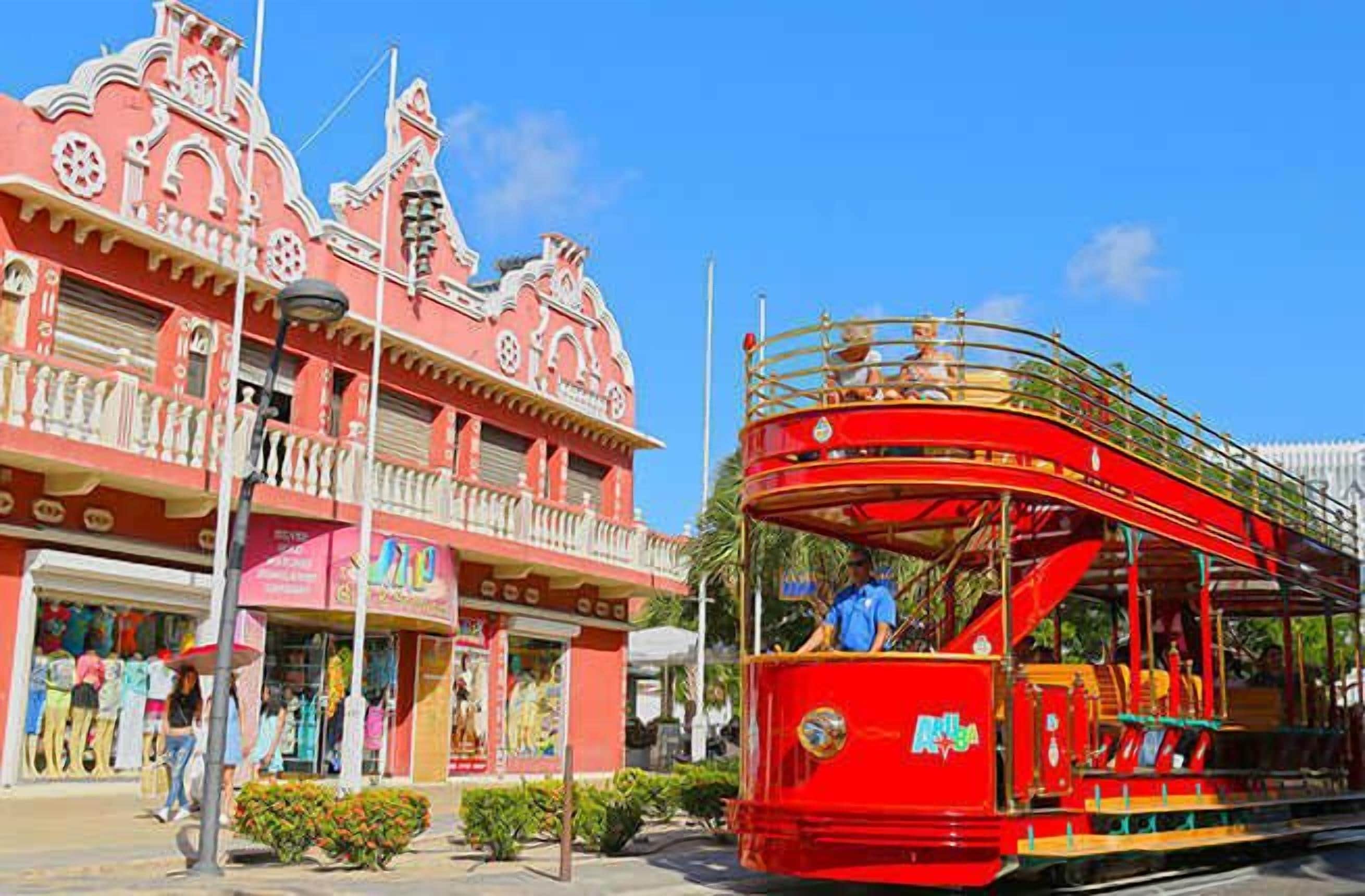 A red double-decker bus with a "Muleba" logo, parked in front of a building with a red facade and white trim.