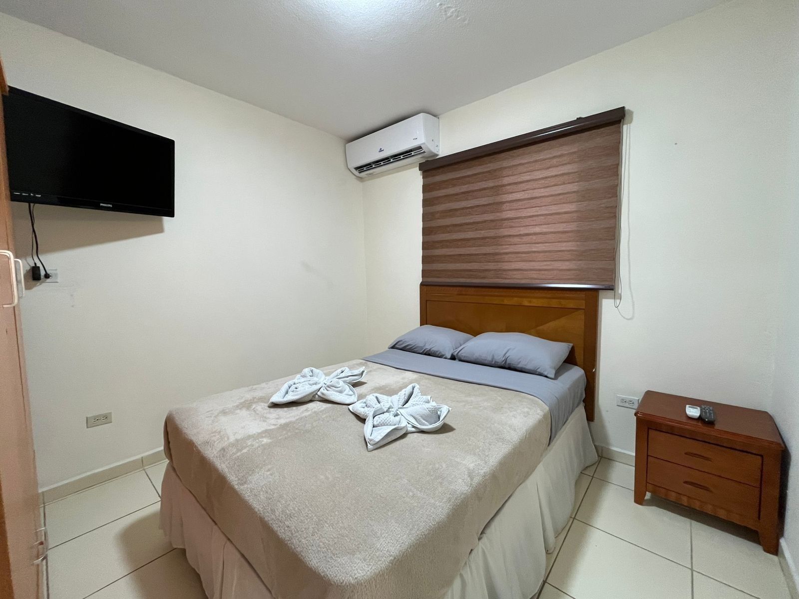 Una camera da letto con un letto, una TV a parete, un condizionatore d'aria, un comodino e un tavolino di fianco al letto.