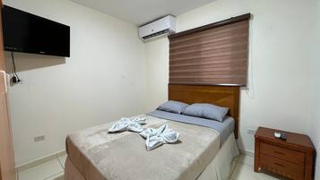 Una camera da letto con un letto, una TV a parete, un condizionatore d'aria, un comodino e un tavolino di fianco al letto.