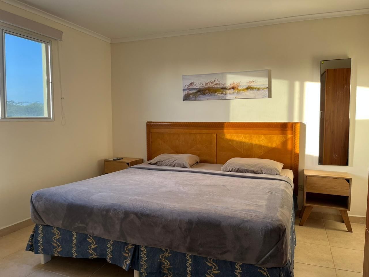 Un dormitorio con cama, mesita de noche, cabecera de madera, un cuadro en la pared y ventana.