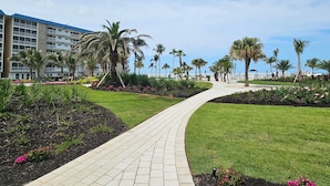 A paved pathway leading through a landscaped area with palm trees and flower beds, flanked by a multi-story hotel building.