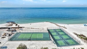 A tennis court complex with multiple courts, a sandy beach, and a view of the ocean.