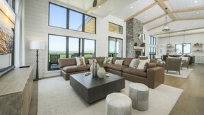 A spacious living area with a large sectional sofa, a coffee table, and a fireplace. There are multiple windows offering views of the outdoors.