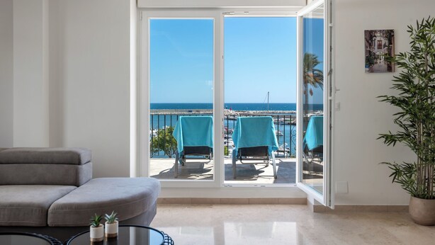 A living room with a large sliding glass door leading to a balcony with a view of the sea and boats. - Belgravia Club Townhouse Estepona by Mabiente Properties (Estepona)