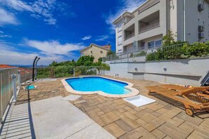 A rooftop swimming pool with a wooden deck and lounge chairs, surrounded by a white building with balconies. - Holiday flat with pool (Crikvenica)