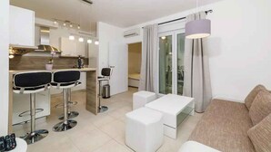 A modern apartment with a kitchen, dining area, and living room. It includes two bar stools, a white sofa, and a glass dining table.