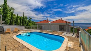 A swimming pool with a tiled deck, lounge chairs, and a view of a red-roofed building and trees.