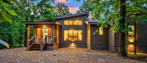 A modern two-story house with a covered porch, a gravel driveway, and a well-lit interior.