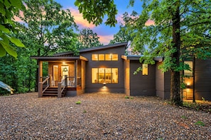 A modern two-story house with a covered porch, a gravel driveway, and a well-lit interior. - Family Escape | Hot Tub + Playset | Sleeps 10 (Broken Bow)