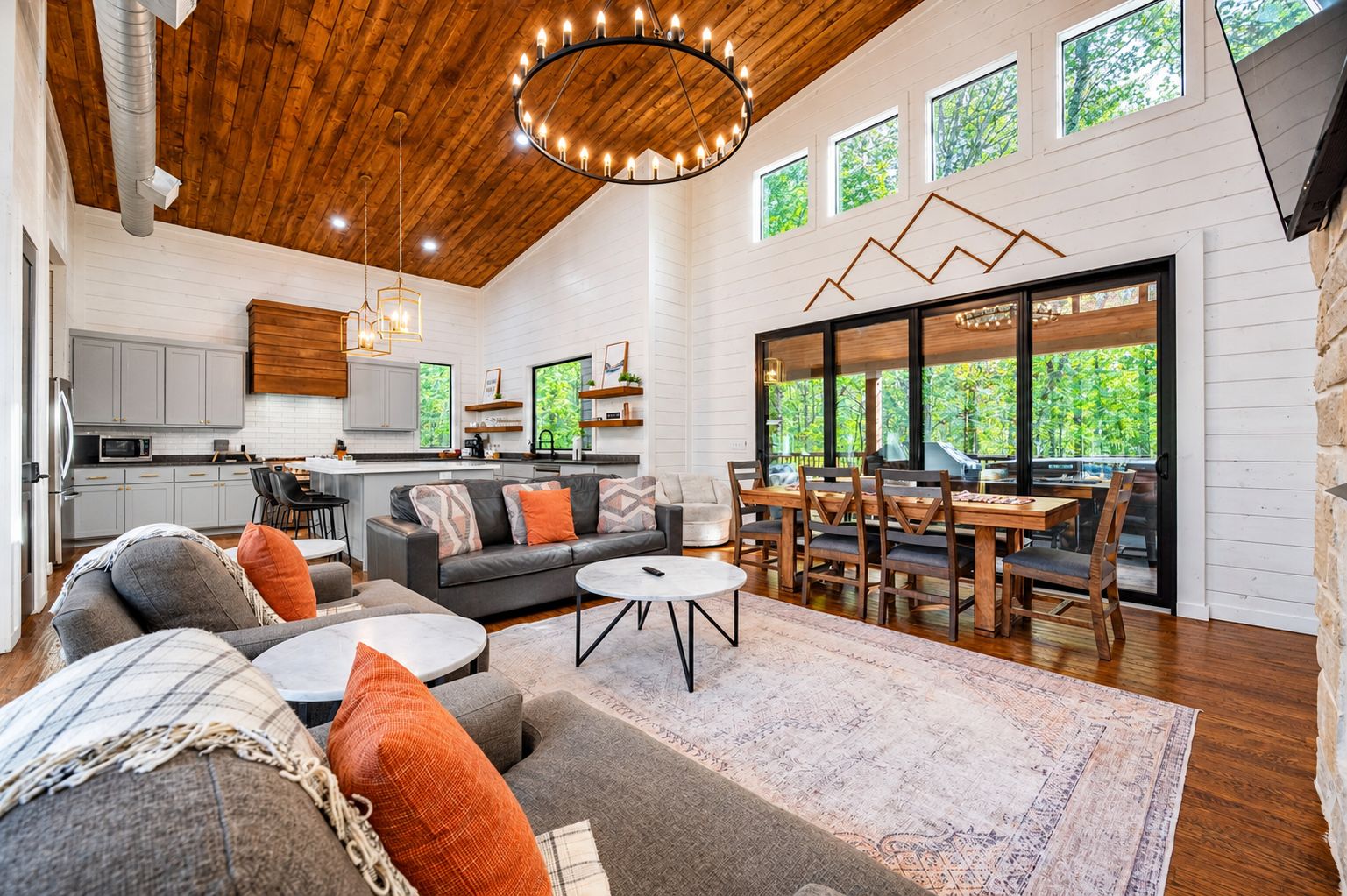 A spacious living area with a wooden ceiling, a dining table with chairs, a sofa set, and a kitchen in the background.