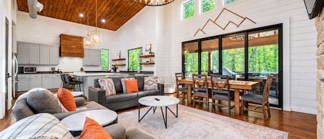 A spacious living area with a wooden ceiling, a dining table with chairs, a sofa set, and a kitchen in the background.