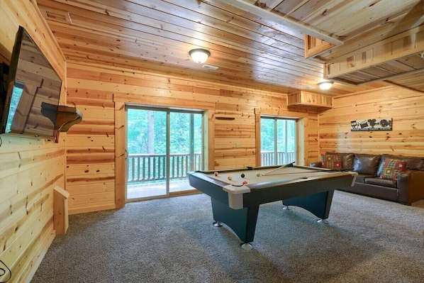 A wooden cabin interior with a pool table, a sofa, and a large window.