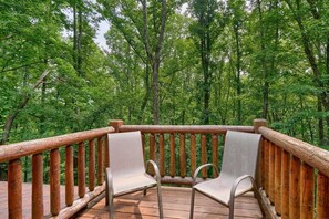 A wooden deck with two metal chairs and a wooden railing overlooking a forest.