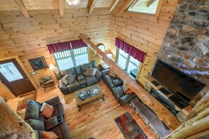 Interior acogedor de una cabaña de troncos con chimenea de piedra, pisos de madera y un ventanal amplio.
