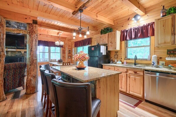 A rustic cabin-style kitchen with wooden cabinets, a granite island, and a stone fireplace. - Tall Timbers Lodge | Hocking Hills, 5BR, Pets (Logan)