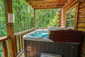 A wooden cabin with a hot tub on a deck surrounded by trees.