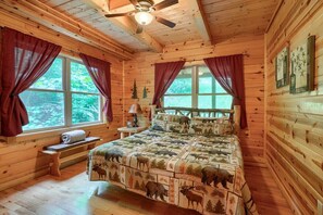 Una habitación acogedora tipo cabaña con interior de madera, una cama con colcha de motivos naturales, un ventilador de techo y una ventana con cortinas.