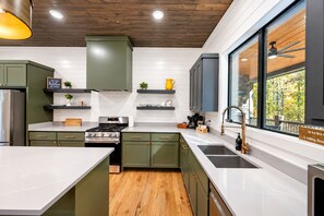 A modern kitchen with green cabinets, a stainless steel stove, and a large window offering a view of trees.