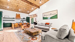 A spacious living area with a wooden ceiling, a gray sofa, a coffee table, and a kitchen in the background. - Play & Stay | 4BR Playset, Pool/Ping Pong, Arcade (Broken Bow)