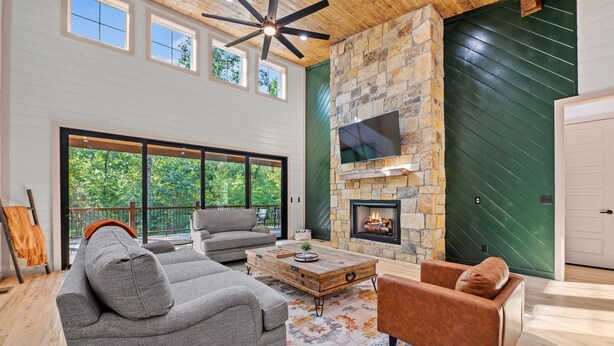 A spacious living room with a stone fireplace, large windows, and a ceiling fan. - Play & Stay | 4BR Playset, Pool/Ping Pong, Arcade (Broken Bow)