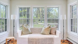 Ein Zimmer mit einem weißen Sofa, zwei Stehlampen und großen Fenstern mit Blick auf Bäume und einen Garten.