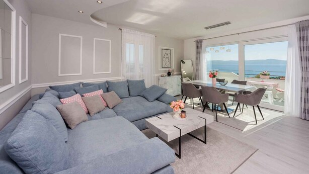 A modern living room with a large grey sectional sofa, a dining area with a table and chairs, and a large window offering a view of the sea. - Holiday apartment with balcony (Crikvenica)