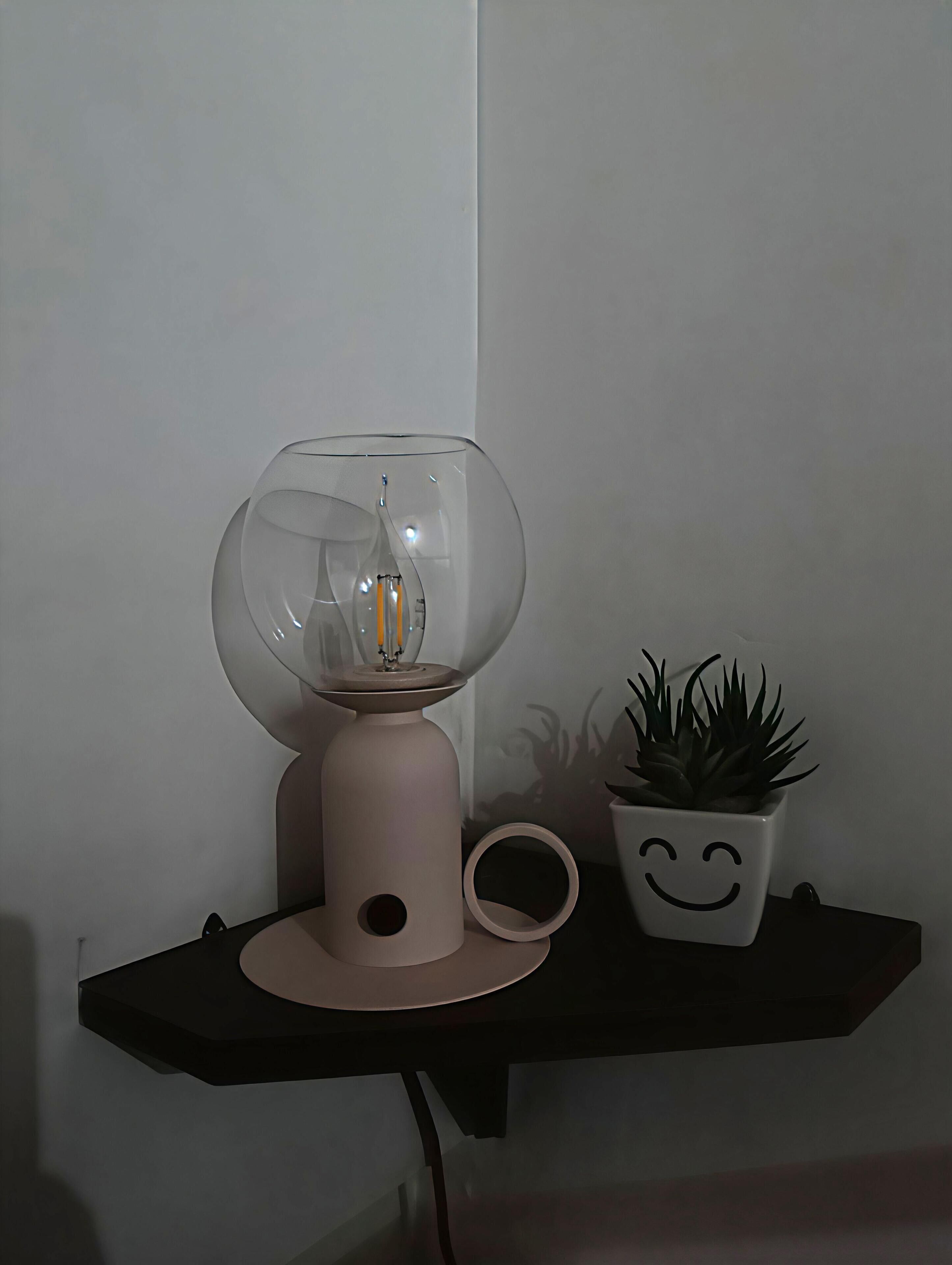 A modern lamp with a clear glass enclosure and a small plant in a smiley face pot on a black shelf.