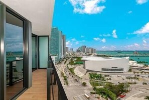 Un bâtiment moderne à la façade blanche et incurvée, doté d’un balcon avec des garde-corps en verre, et offrant une vue sur un paysage urbain avec de hauts immeubles et un ciel bleu dégagé.