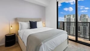 Quarto moderno com uma cama grande, uma mesa de cabeceira e vista para a cidade através de janelas do chão ao teto.