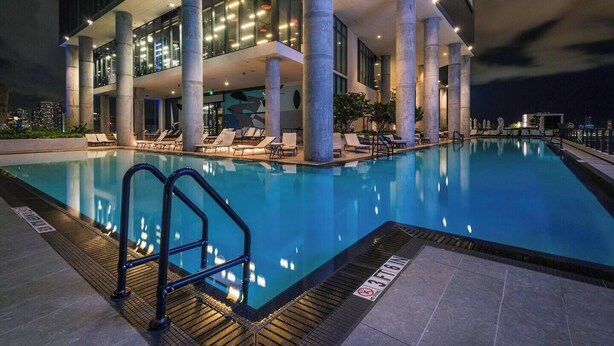 Un área de piscina moderna en un hotel, con vista despejada a la piscina, sillas de descanso y un edificio grande de varios pisos y columnas.