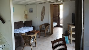 Un espace de vie compact comprenant un canapé, une petite table, un fauteuil en osier, un lit et un téléviseur.
