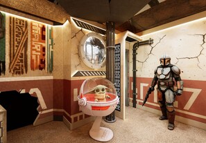 Een kamer met een Star Wars thema, inclusief een levensgroot Bounty Hunter figuur, een Baby Yoda figuur in een transparante cocon, en een muur met een gescheurde textuur.