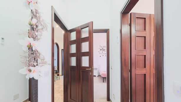 A hallway with wooden doors, a white wall with a decorative floral arrangement, and a bed with a pink blanket in the background. - The prestigious corner of Forli (Forlì)