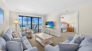 Un vaste salon comprenant un grand canapé, une table basse et un téléviseur posé sur un meuble. Un balcon offre une vue sur l’océan et les palmiers.