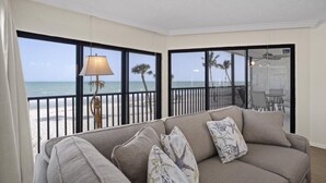 A balcony with a view of the beach and ocean, a sofa with decorative pillows, and a table with chairs.