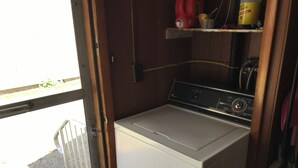 A compact laundry room with a front-loading washer and dryer, a small shelf with cleaning supplies, and a window providing a view of the outside. - Quaint Village home in Allegany.1 mile from St. Bonaventure. (Allegany)