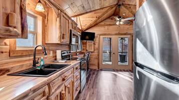 A wooden cabin interior with a kitchen featuring a stainless steel refrigerator, wooden cabinets, and a large window.