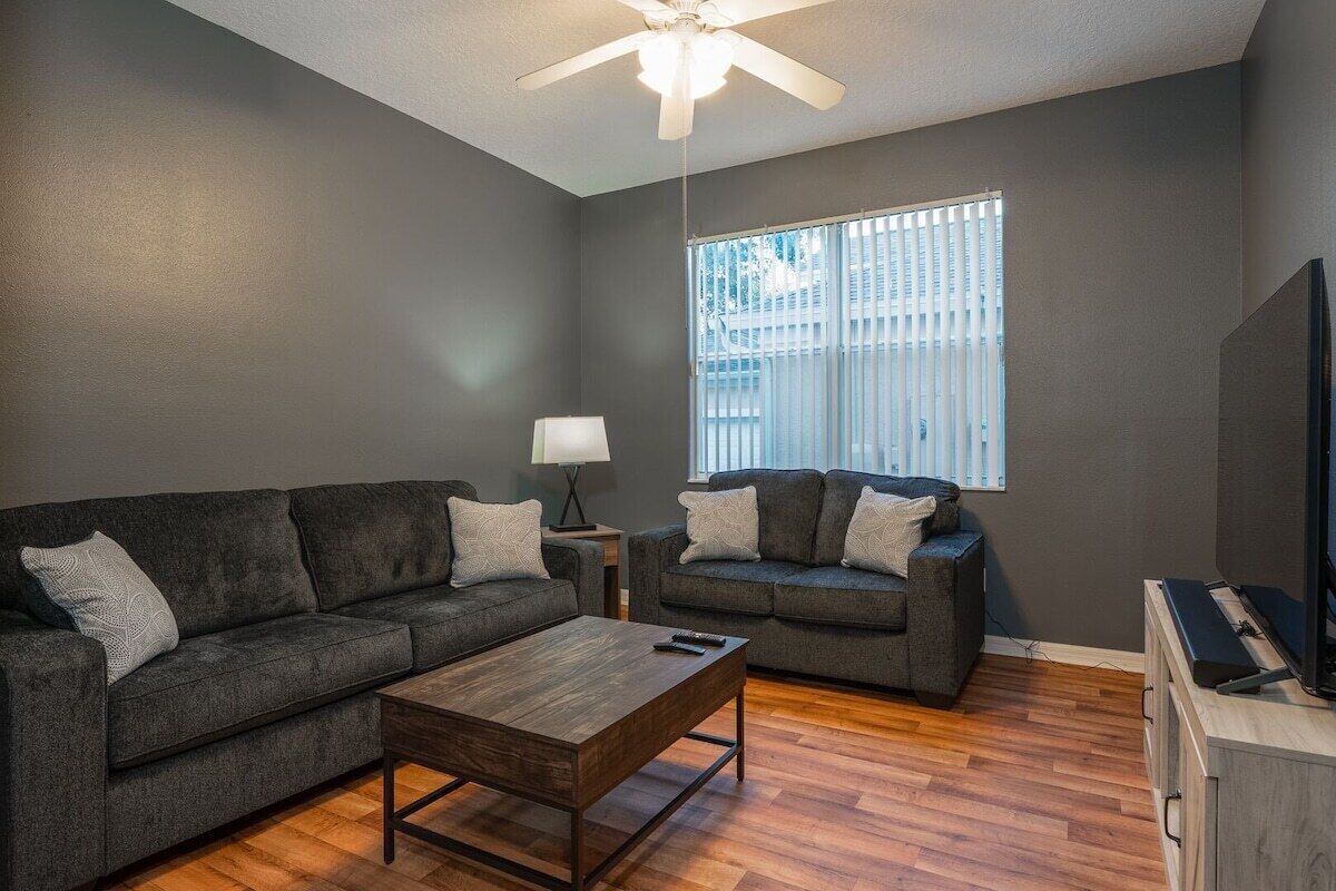 A living room with a dark gray wall, a ceiling fan, a large window with blinds, a sofa, a coffee table, and a TV on a wooden cabinet.