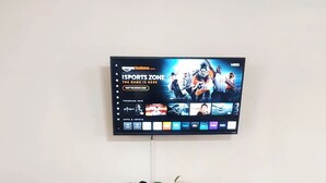 A flat-screen TV mounted on a wall displaying a sports channel. In front of the TV, there is a wooden console table with a white vase containing flowers and two candles.