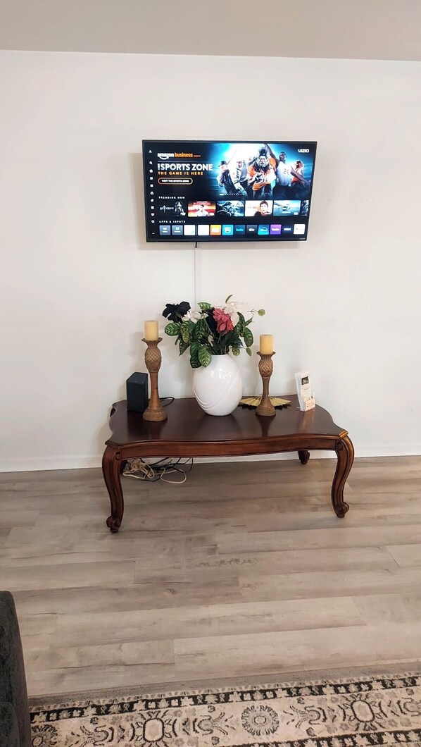 En platt-TV är monterad på väggen och visar en sportkanal. Framför TV:n står ett konsolbord i trä med en vit vas som innehåller blommor och två ljus.