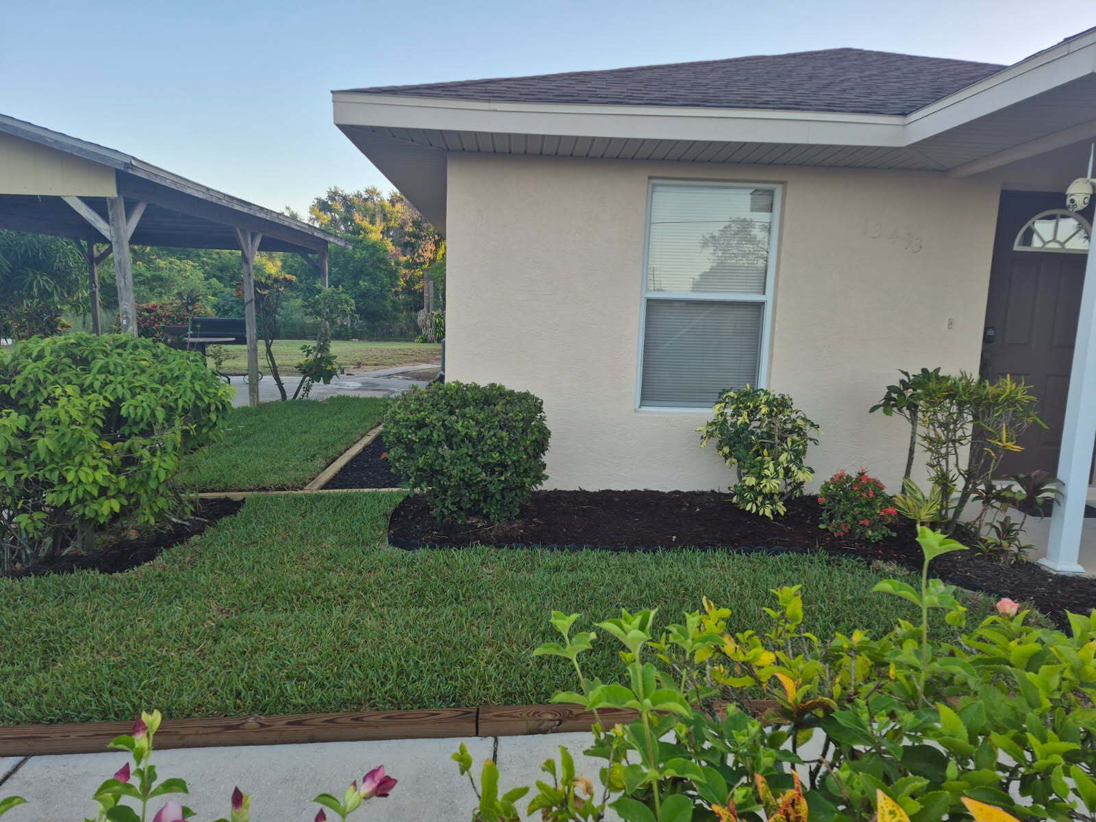 A residential house with a well-maintained lawn, a small garden area with various plants, and a clear sky.
