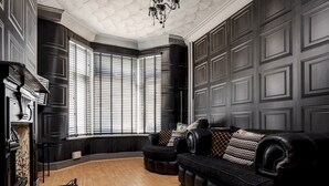 Una habitación con piso de madera oscura, un sofá de cuero negro con cojines estampados, una chimenea y un ventanal con persianas.